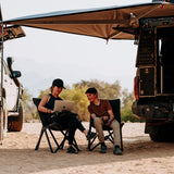 Two people sitting under a vehicle mounted an Alu-Cab 270 Shadow Awning, one using a laptop. Camp4x4. camp 4x4, overlanding, camping. Double Cab. Single Cab. Extra Cab