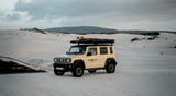 Suzuki Jimny 4x4 with surfboard and Alu-Cab Rooftop pop up tent driving on sand dunes.
