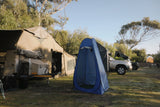 Camping scene with a tent, trailer, and blue pop-up shower cubicle tent in a grassy area with trees.
