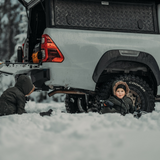 Two children playing in the snow next to a white truck with an Alu-cab Explorer Canopy, with treaded finish, and visible 'Cooper' tire brand. Camp4x4 camp 4x4, overlanding, camping