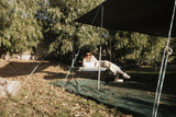 Person sitting on a stretcher mattress under a tarp in a natural setting with a laptop.